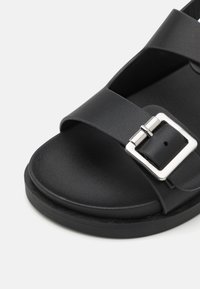 Sandálias pretas com duas tiras largas e uma fivela quadrada prateada. Material em pele sintética lisa, bico arredondado e sola texturizada.