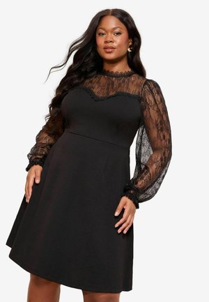 Kvinde iført en sort kjole med gennemsigtige blonder lange ærmer og høj hals, poserer med hænderne tæt på hoften mod en hvid baggrund.