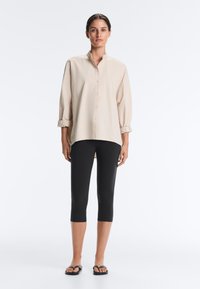 Woman with dark hair wearing a beige button-up shirt with rolled sleeves, black cropped leggings, and black sandals, standing against white background.