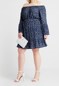 Avskuren ärm marinblå klänning med ormtryck, volangnederkant och åtdragen midja; kombinerad med en vit handväska och remsandaler.