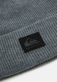 Pletená šedá čepice s ribby texturou a černou koženou nášivkou s logem. Design nemá žádné další vzory nebo akcenty.