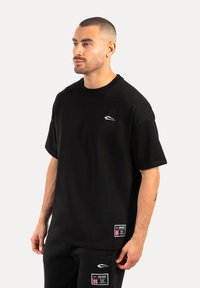 Camiseta de algodón negra con mangas cortas, escote redondo y pequeño logo blanco en el pecho. Presenta dos etiquetas en el dobladillo inferior.