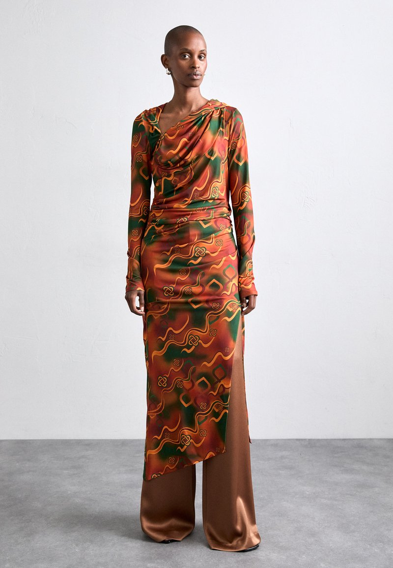 Long-sleeved, draped dress made of smooth green and orange patterned fabric, featuring a high slit, paired with brown satin pants.