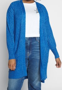 Cardigan à manches longues et à ouverture devant de couleur bleu vif, avec une texture côtelée et deux poches latérales, superposé à un t-shirt blanc et un jean.
