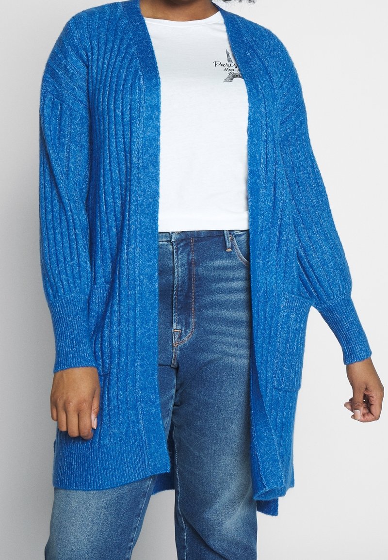 Cardigan à manches longues et à ouverture devant de couleur bleu vif, avec une texture côtelée et deux poches latérales, superposé à un t-shirt blanc et un jean.
