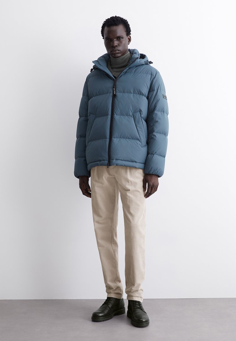 Gepolsterte türkisfarbene Jacke mit Kapuze und Reißverschluss, mit einem gesteppten Design. Getragen über einem grünen Rollkragenpullover, kombiniert mit hellbeigen Hosen und grünen Stiefeln.