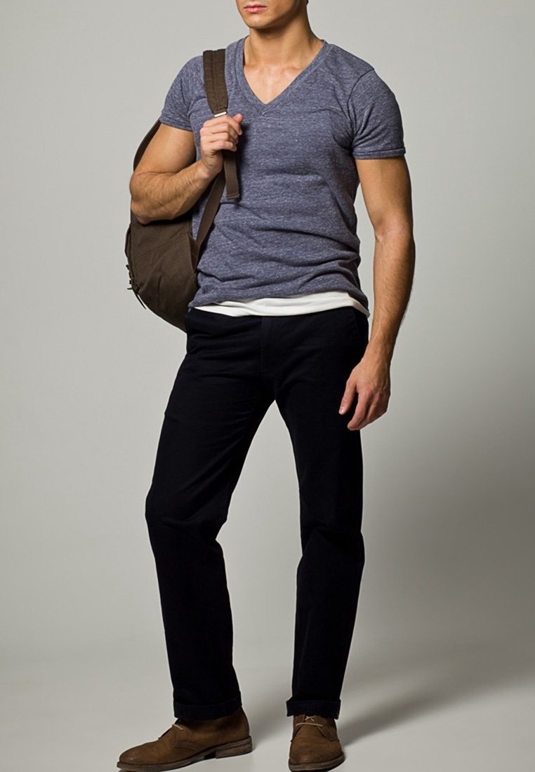 Homme portant un t-shirt gris à col en V, pantalon noir, chaussures marron, portant un sac à épaule marron sur un fond uni.