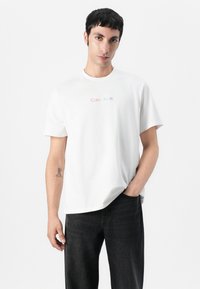 Witte katoenen t-shirt met korte mouwen, voorzien van een meerkleurig "Calvin Klein" logo op de borst. Gecombineerd met donkere jeans.