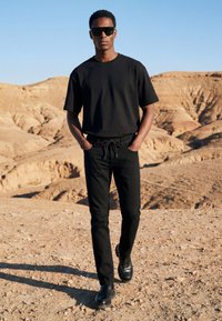 Homme portant des lunettes de soleil noires, un t-shirt noir, un pantalon noir et des chaussures noires, debout sur un paysage désertique rocheux sous un ciel clair.