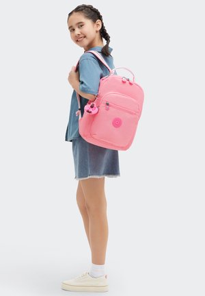 Rosa ryggsäck med rundad form, tygmaterial, framficka med dragkedja och dekorativ charm. Bärs av ett barn med flätat hår.