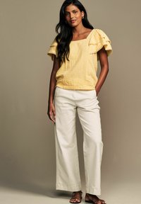 Top giallo, texture con maniche arricciate, scollo quadrato; abbinato a pantaloni bianchi a gamba larga e sandali marroni con cinturini.