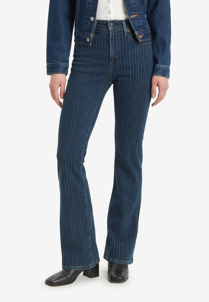 Jean en denim bleu évasé avec des rayures verticales blanches, taille haute et un design classique à cinq poches. Porté avec des bottines noires à la cheville.