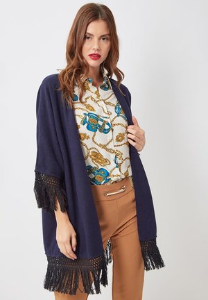 Mujer con cárdigan abierto azul marino con flecos sobre blusa blanca con estampado de cadenas azules y doradas y pantalones marrones con detalle de cinturón.