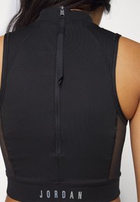 Detailní záběr na černý sportovní top bez rukávů se zipem na zadní straně a nápisem "JORDAN" vytištěným na spodním lemu.