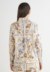Chemise à motifs floraux beige, ornée d'accents bleus et marron. Manches longues avec col classique et coupe décontractée, affichant un détail de poche.