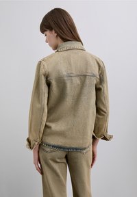 Jachetă din denim maro deschis cu croială relaxată, detalii vizibile de cusătură și manșete răsucite. Purtați cu pantaloni asortați maro deschis.