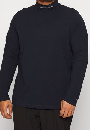 Mężczyzna w czarnym, długim rękawem, golfie z logo "Tommy Hilfiger" na kołnierzu i małym logo flagi na mankiecie lewej rękaw.
