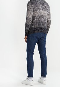 Gestrickter Pullover mit einem Farbverlaufsmuster von hellem Beige bis dunklem Marineblau, kombiniert mit dunkelblauen Jeans und weißen Turnschuhen.
