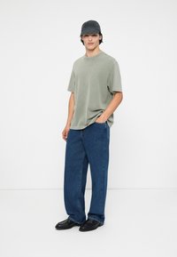 Svetlozelené bavlnené tričko, vo voľnom strihu, skombinované s širokými modrými džínsami. Čierna čiapka s logom a čierne topánky dotvárajú outfit.