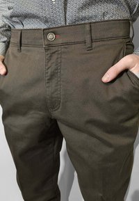Pantaloni verde oliva scuro con un motivo a trama fine, dotati di tasche laterali e chiusura con bottone, abbinati a una camicia a fantasia.