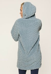 Manteau à capuche, ouvert à l'avant, en polaire texturée de couleur bleu clair avec un motif en écailles. Il a des manches courtes et une ourlet finissant au-dessus du genou.