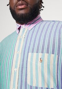 Camisa de rayas multicolores con tonos verdes, azules y amarillos. Tiene un bolsillo con un logo bordado y un cuello rosa.