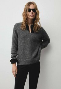 Maglione grigio lavorato a maglia con spruzzi argentati, scollatura a V e polsini neri, abbinato a pantaloni neri. Presenta una vestibilità comoda e una texture morbida.