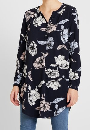 Frau trägt eine langärmelige schwarze Tunika mit großen weißen und hellrosa Blumenmustern, kombiniert mit blauen Jeans.