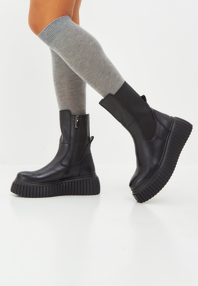 Botas de tobillo de imitación de cuero negro con cierre de cremallera y suelas gruesas acanaladas. Combinadas con calcetines grises hasta la rodilla. Textura suave, diseño moderno.