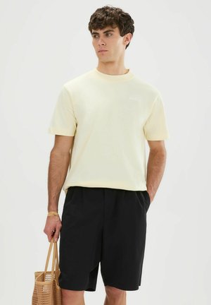 Joven con una camisa de manga corta amarillo pálido y pantalones cortos negros, sosteniendo una bolsa de mano marrón tejida, mirando hacia un lado.