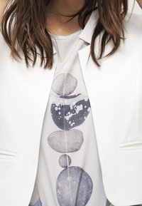 Blazer blanco a medida sobre una blusa clara que presenta un patrón vertical de acuarela con piedras redondeadas en tonos grises y azules, con detalles de salpicaduras.