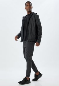 Sort letvægtsjakke med hætte, sort T-shirt, grå tapered joggers og sorte sneakers, som går på en ensfarvet baggrund.