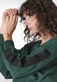 Zelená bavlněná mikina s bočním designem, který zahrnuje texturovanou černou látku a vyražené písmo na rukávě; kulatý výstřih a volnější střih.