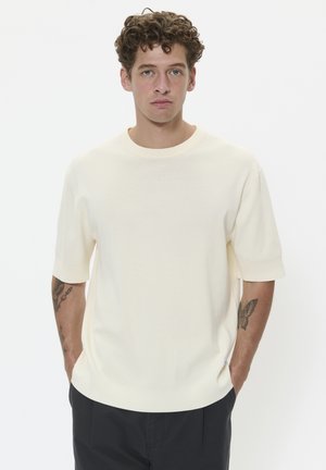 Gros pull à manches courtes de couleur crème claire, avec un col rond, une texture douce et une coupe décontractée, associé à un pantalon sombre. Présence d'un tatouage visible sur le bras.