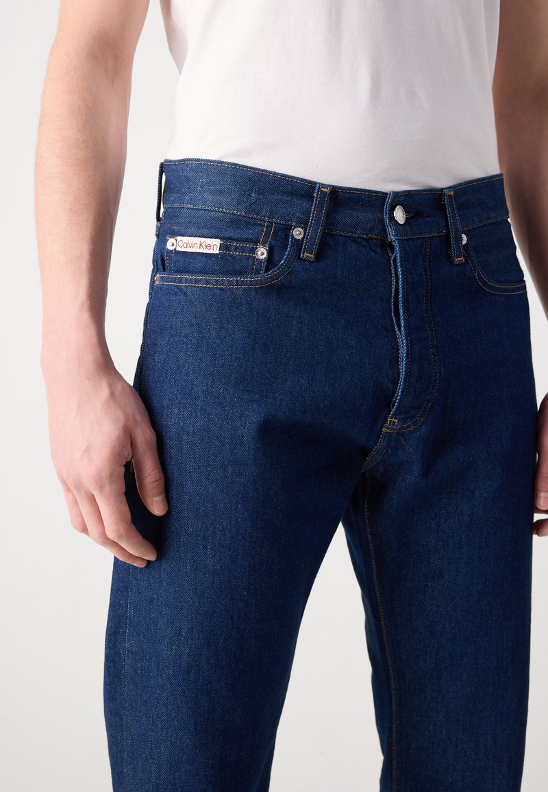 Ciemnobłękitne dżinsy o dopasowanym kroju. Posiadają logo Calvin Klein na pasku, srebrne elementy metalowe oraz klasyczne wykończenia szwów.