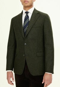 Dunkelgrüner Tweed-Blazer mit Fischgrätenmuster, zwei Knöpfen, Reverskragen und einer Brusttasche. Kombiniert mit einer gestreiften Krawatte und einem weißen Hemd.