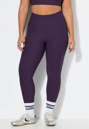 Persona que lleva leggings morados de tiro alto, calcetines blancos con texto negro y zapatillas plateadas con cordones, de pie sobre un fondo blanco.