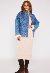 Giacca blu trapuntata con polsini e colletto in shearling, abbinata a un vestito lavorato a maglia beige e stivali marroni al ginocchio.