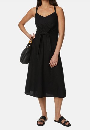 Vrouw in een zwarte mouwloze midijurk met een strik aan de voorkant, zwarte slippers en een zwarte geweven handtas met handvatten van schildpadmotief.