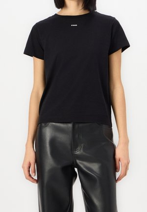 Persona che indossa una maglietta nera a maniche corte con la scritta "PINKO" sul petto e pantaloni di pelle neri, in piedi contro uno sfondo semplice.