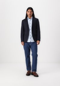 Dark navy blazer layered over a light blue button-up shirt, paired with blue denim jeans and brown dress shoes. Smooth fabric and tailored fit.
