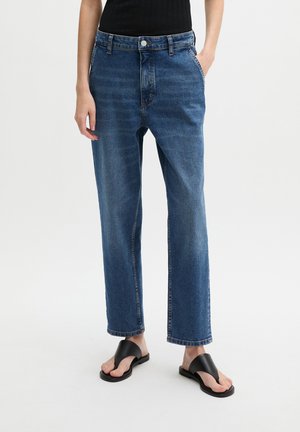 Persoon die blauwe jeans met rechte pijpen, een zwart mouwloos topje en zwarte sandalen met open tenen draagt, staand met één hand in de zak op een witte achtergrond.