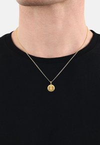 Guldtonad hängande halsband med en cirkulär berlock med ingraverad design, hängande på en tunn kedja, bärs över en svart skjorta.