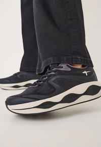Black athletic shoes with textured mesh panels, a cushioned sole, and a wavy midsole design. Lace-up closure with subtle branding detail.