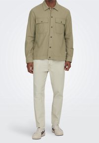 Licht olijfgroene shirtjack met twee borstzakken, knoopsluiting en lange mouwen; crèmekleurige broek en witte sneakers.