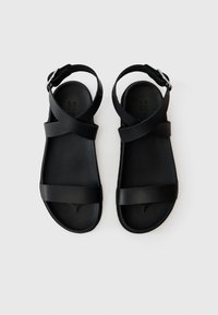 Sorte sandaler med flat såle, med to brede stropper som krysser foran og en justerbar ankelstropp for sikker passform. Glatt tekstur.
