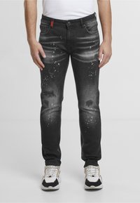Homme portant un jean noir délavé usé avec des éclaboussures de peinture, associé à des baskets blanches et noires, debout devant un fond uni.