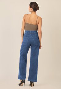 Blaue hochtaillierte weite Jeans mit zwei Gesäßtaschen und einer glatten Denim-Oberfläche, kombiniert mit einem braunen enganliegenden Oberteil und schwarzen Absätzen.