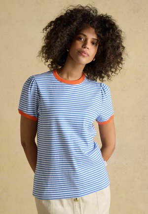 Woman with curly hair wearing a blue and white striped shirt with orange trim, standing against a beige background.
