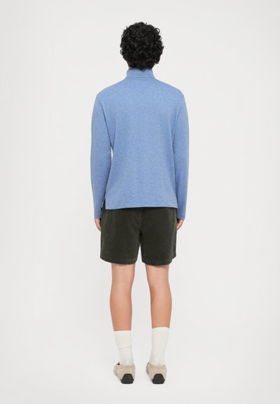 Top azul de manga longa com gola alta, combinado com calções de veludo cotelê verde escuro. O tecido parece suave com uma textura subtil.